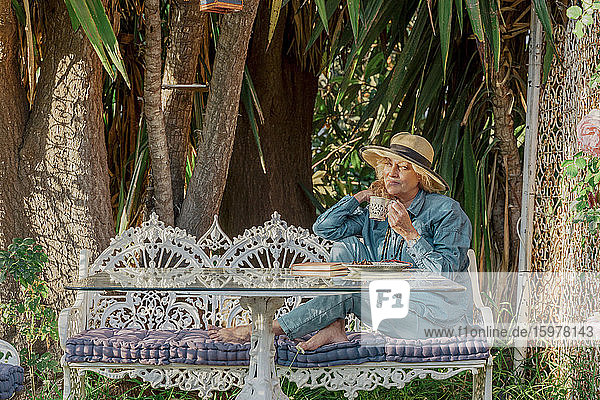 Ältere Frau sitzt auf einer Gartenbank und trinkt eine Tasse Tee