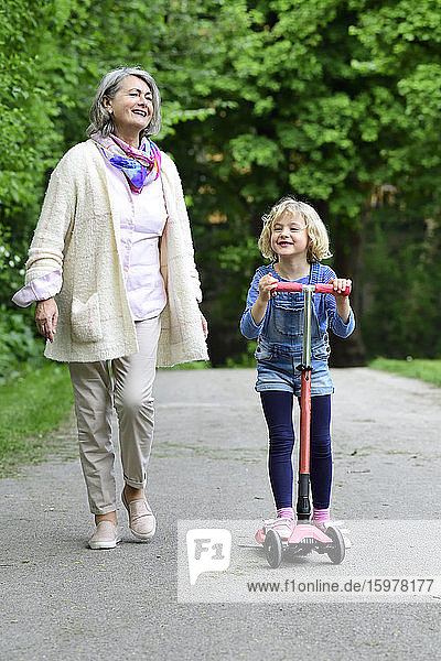 Happy senior woman walking with granddaughter riding push scooter in park
