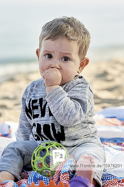 Niedlicher Junge lutscht am Daumen  während er auf einer Decke am Strand sitzt