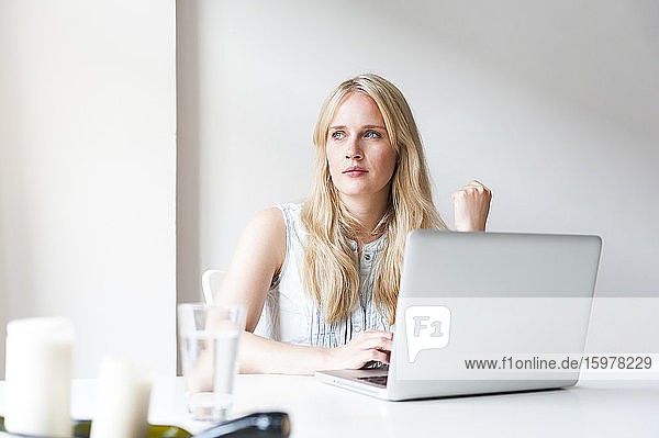 Porträt einer blonden jungen Frau  die mit einem Laptop am Tisch sitzt