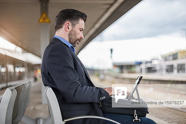 Seitenansicht eines selbstbewussten reifen Geschäftsmannes  der an einem digitalen Tablet arbeitet  während er am Bahnhof wartet