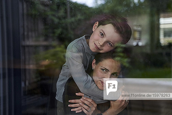 Liebender Sohn  der seine Mutter umarmt  während er zu Hause steht  gesehen durch ein Fenster