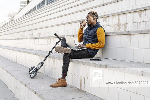 Junger Mann mit Laptop und Motorroller sitzt auf einer Treppe und trinkt Kaffee
