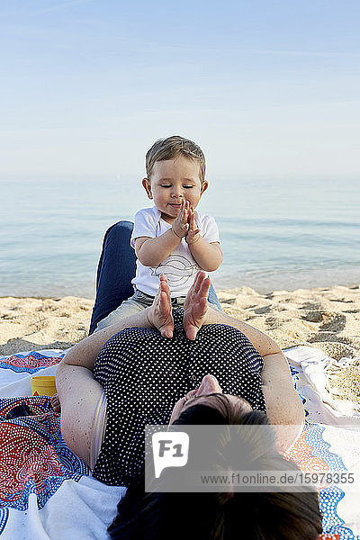 Mother playing clapping game with cute son while lying on blanket at beach against sea