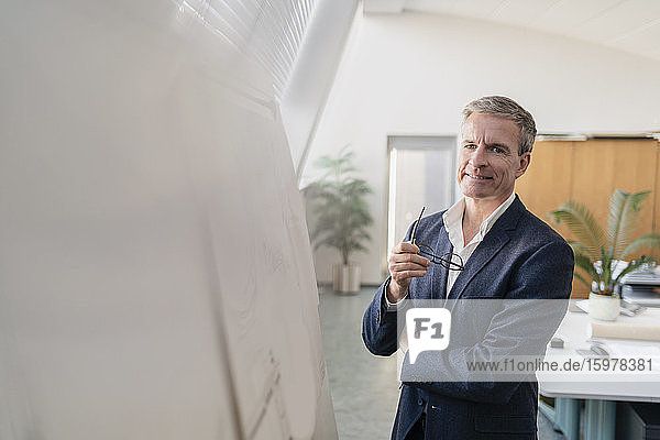 Porträt eines lächelnden männlichen Unternehmers  der eine Brille hält und an einem Whiteboard im Büro steht