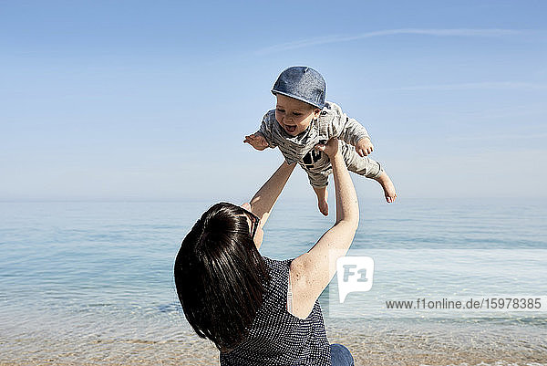 Mother picking up happy cute son against sea during sunny day