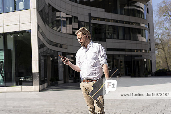 Geschäftsmann hält Skateboard und überprüft Smartphone in der Stadt