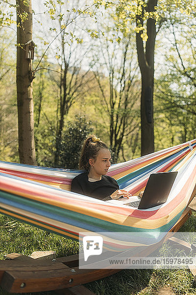 Junge Frau  die in einer Hängematte im Garten sitzt und am Laptop arbeitet