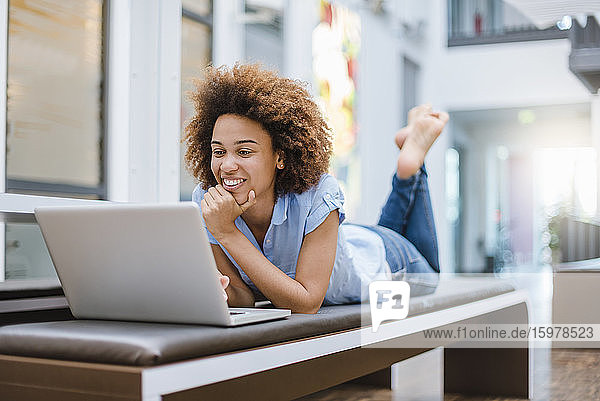 Junge Frau liegt auf einer Bank in einem modernen Büro und benutzt einen Laptop