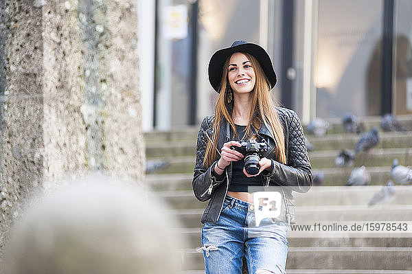Happy young woman holding DSLR camera on steps in city