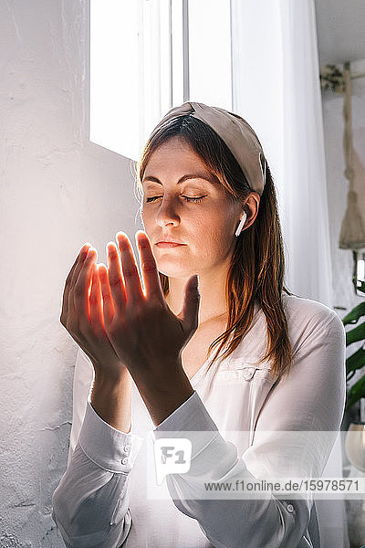 Schöne Frau betet mit geschlossenen Augen am Fenster zu Hause