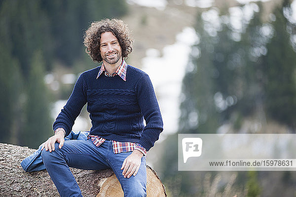 Lächelnder gut aussehender Mann  der auf einem Baumstamm sitzt und wegschaut