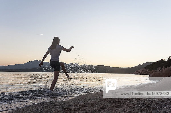 Rückenansicht einer Frau  die bei Sonnenuntergang am Meeresufer im Wasser plantscht  Sardinien  Italien