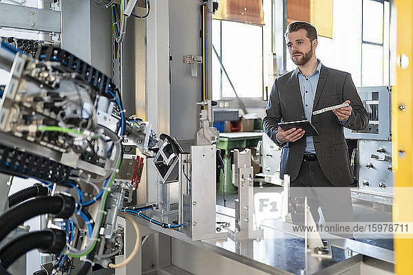 Young businessman with tablet and workpiece at a machine in a factory