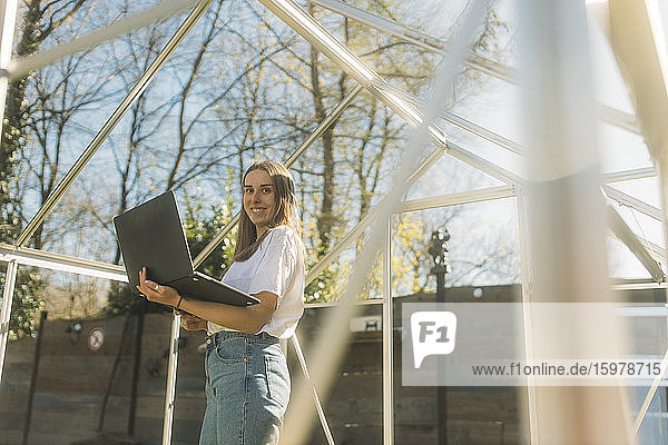 Lächelnde Frau  die einen Laptop hält  während sie in einem unvollständigen Gewächshaus im Hof steht