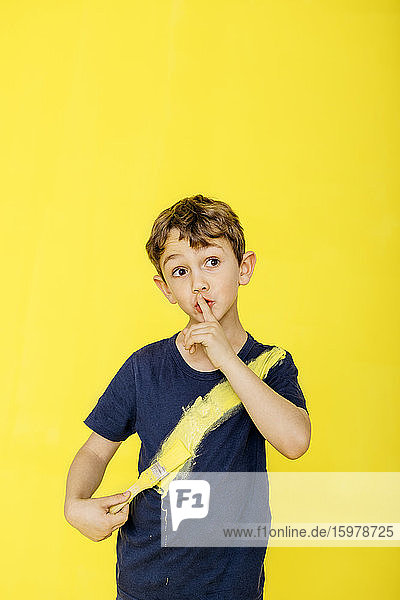 Süßer Junge mit Pinsel auf T-Shirt  während er mit dem Finger auf den Lippen vor gelbem Hintergrund steht