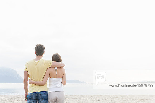 Rückenansicht eines Paares  das Arm in Arm am Strand steht und auf das Meer blickt  Sardinien  Italien