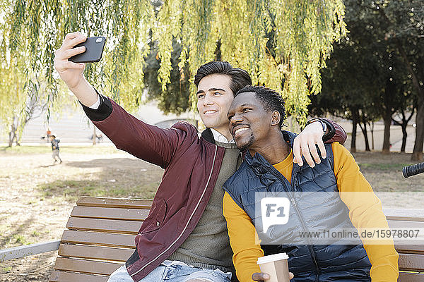 Zwei glückliche junge Männer sitzen auf einer Parkbank und machen ein Selfie
