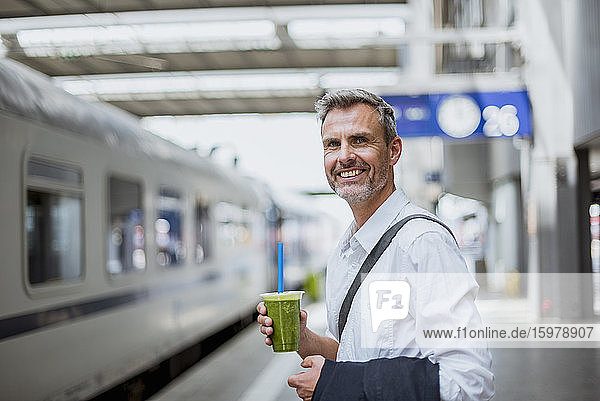 Lächelnder Geschäftsmann hält ein Getränk in der Hand  während er am Bahnsteig steht