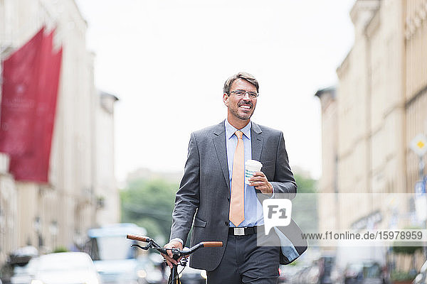 Lächelnder Geschäftsmann mit Kaffee in der Hand  der wegschaut  während er mit dem Fahrrad in der Stadt spazieren geht