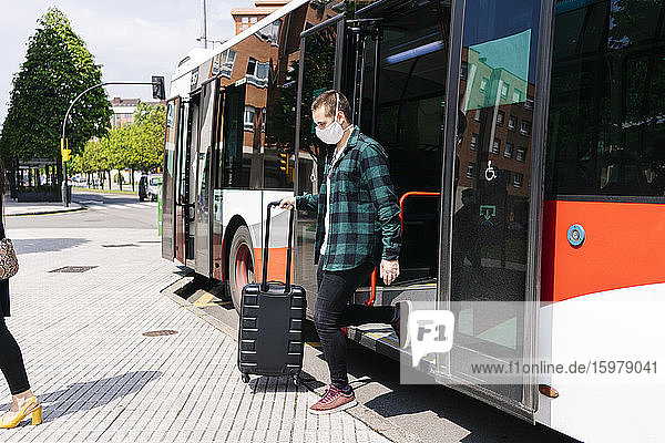 Young man wearing protective mask getting off public bus with trolley bag  Spain