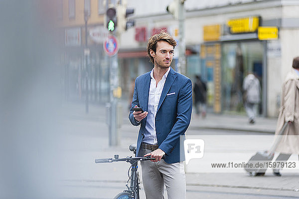 Porträt eines jungen Geschäftsmannes mit Fahrrad und Smartphone  der in der Stadt wartet