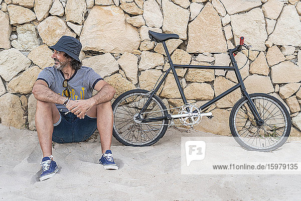 Älterer Mann mit Fahrrad sitzt an der Wand am Strand