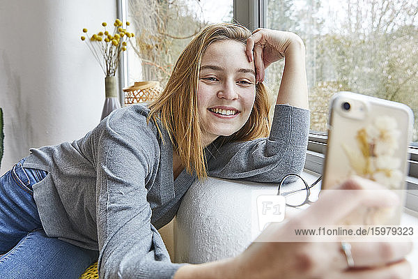 Porträt einer lächelnden jungen Frau  die sich zu Hause auf die Fensterbank lehnt und ein Selfie mit ihrem Smartphone macht