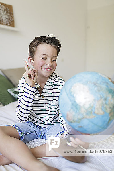 Porträt eines lächelnden kleinen Jungen auf dem Bett sitzend mit seinem Globus