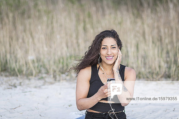 Porträt einer lächelnden jungen Frau am Strand mit Smartphone und Kopfhörern