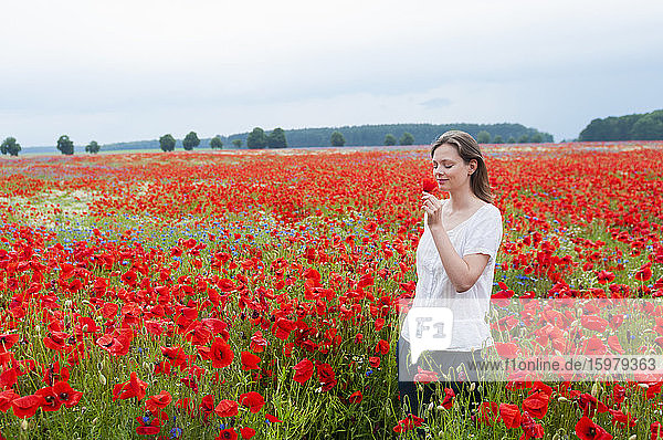 Schöne Frau riecht an Mohn  während sie inmitten eines rot blühenden Feldes gegen den Himmel läuft