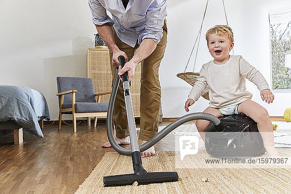 Niedriger Ausschnitt eines Mannes  der einen Teppich reinigt  während ein süßer kleiner Junge auf dem Staubsauger im Wohnzimmer sitzt