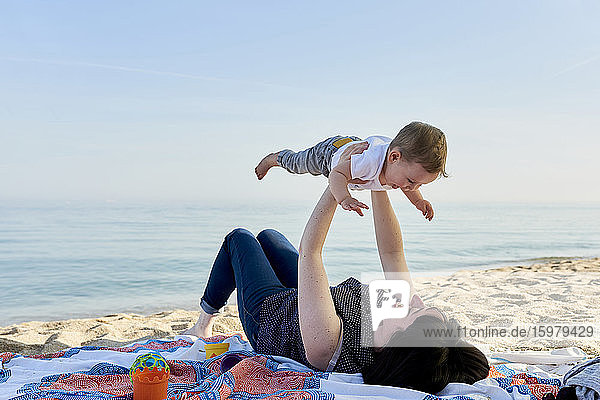 Mother picking up happy son while lying on blanket at beach against sea