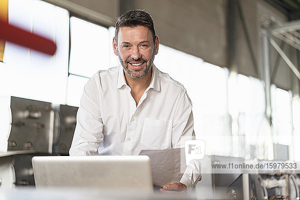 Porträt eines lächelnden reifen Geschäftsmannes  der Papiere hält und einen Laptop in einer Fabrik benutzt