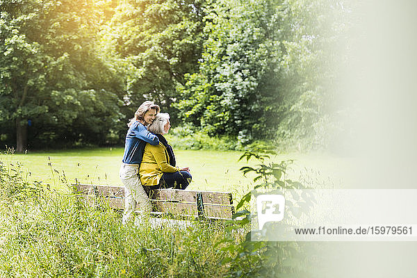 Erwachsene Tochter  die ihre Mutter umarmt  die auf der Rückenlehne einer Parkbank sitzt