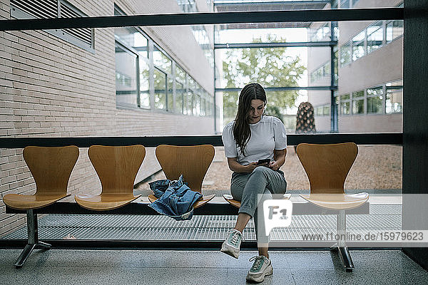 Junge Studentin in voller Länge  die ein Smartphone benutzt  während sie im Korridor einer Universität sitzt