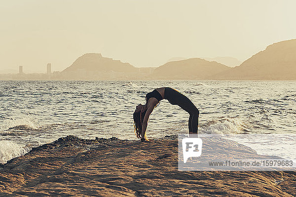 Mature woman practicing yoga at rocky beach in the evening  bow pose