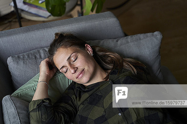 Lächelnde erwachsene Frau mit geschlossenen Augen  die sich auf dem Sofa im Wohnzimmer entspannt