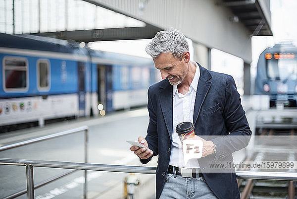 Geschäftsmann mit Kaffee in der Hand und Smartphone im Bahnhofsbereich