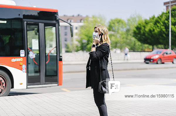 Ältere Frau am Telefon mit Schutzmaske wartet an der Bushaltestelle  Spanien