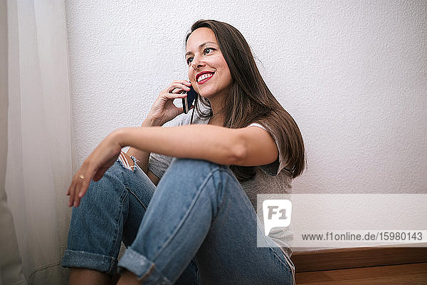 Glückliche Frau  die an einer weißen Wand sitzt und zu Hause mit ihrem Smartphone telefoniert