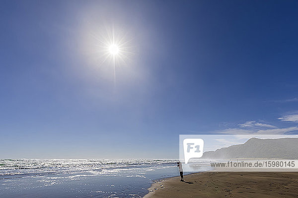 Silhouette einer weiblichen Touristin beim Fotografieren am Piha Beach vor blauem Himmel  Auckland  Neuseeland