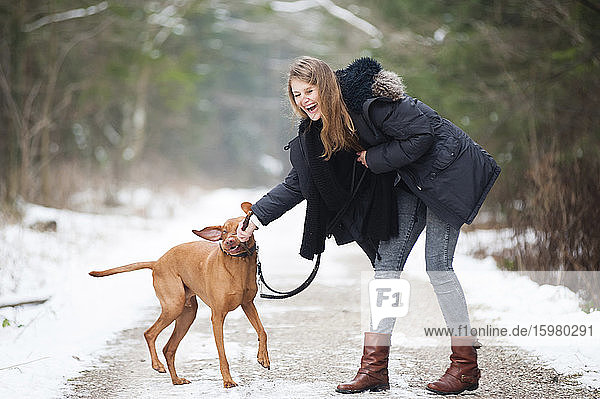 Cheerful young woman playing with dog on road in forest during winter