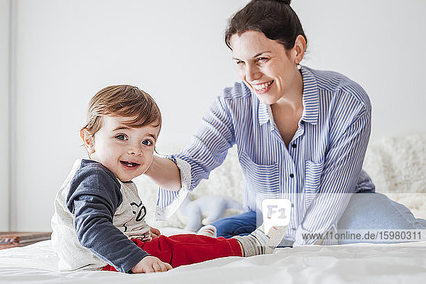 Porträt eines kleinen Jungen auf dem Bett sitzend mit seiner Mutter im Hintergrund