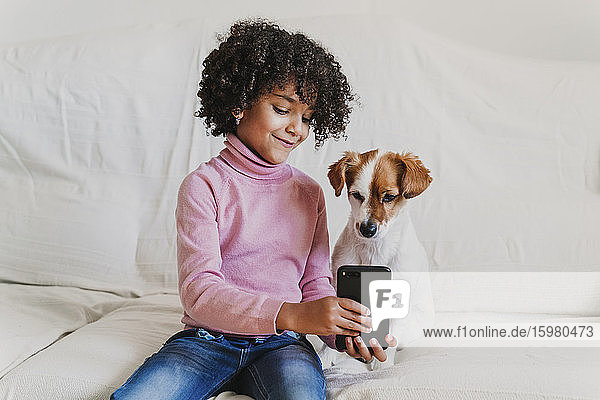Portrait of smiling little girl sitting on the couch with her dog taking selfie with smartphone