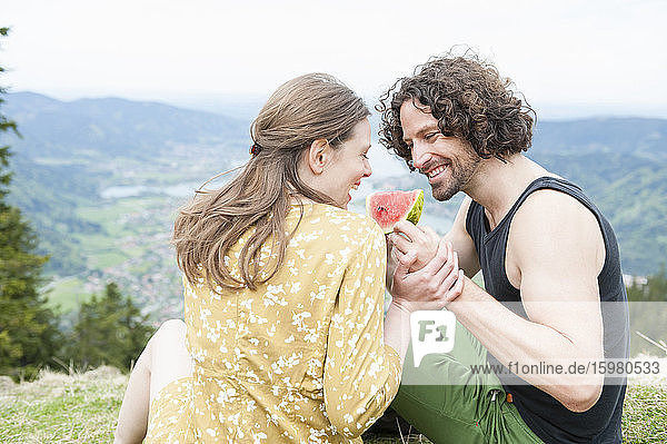 Glückliches erwachsenes Paar  das auf einem Berggipfel sitzend ein Stück Wassermelone genießt
