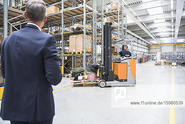 Businessman and worker with forklift in high rack warehouse