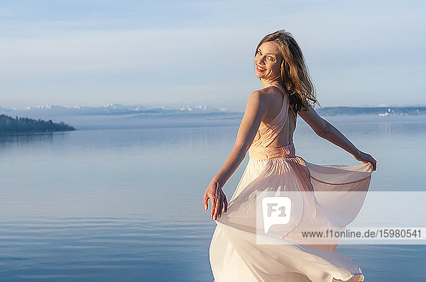 Rückansicht Porträt einer lächelnden schönen Frau im Kleid am Seeufer an einem sonnigen Tag stehend
