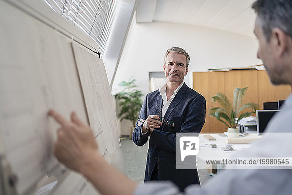 Selbstbewusster reifer Geschäftsmann  der einem männlichen Kollegen zuhört  der während einer Besprechung im Büro eine Strategie am Whiteboard erläutert