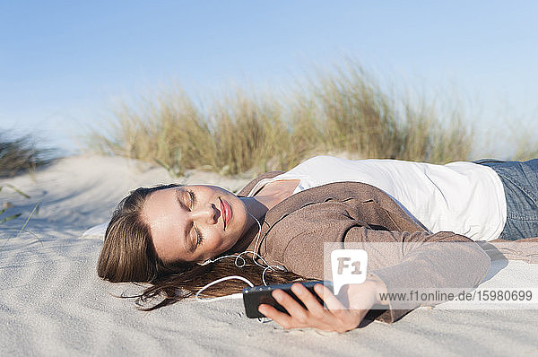 Porträt einer auf einer Stranddüne liegenden Frau  die mit Kopfhörern und Smartphone Musik hört  Sardinien  Italien